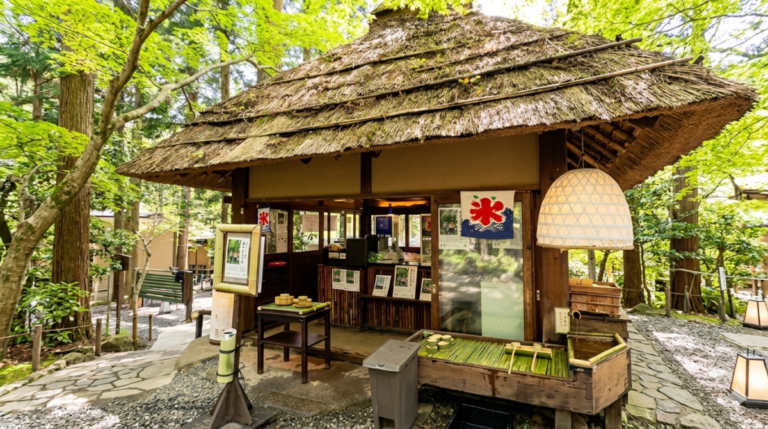 新緑の美しい森の中に佇む、日本の伝統的な茅葺き屋根の茶屋。店頭にはかき氷の旗（氷旗）や竹製の水盤、柄杓が置かれた風情ある和風の景観。