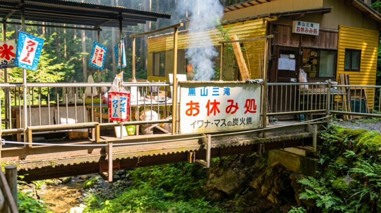 黒山三滝お休み処埼玉県越生町の黒山三滝近くにある休憩所「黒山三滝お休み処」の外観。豊かな自然の緑と清流に囲まれた小さな橋の奥に黄色い木造の建物があり、川魚の炭火焼きの煙が立ち上っています。「イワナ・マスの炭火焼」や「アイスクリーム」の看板が見えます。